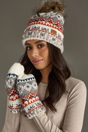 A woman in a beige long-sleeve top wears the Panache Apparel Collection Desi Pom Hat with its faux fur pom and matching gray, white, orange, and blue mittens, smiling softly against a neutral background.