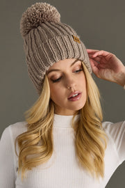 Woman wearing a taupe knitted beanie with a pom-pom against a neutral background