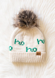 Beige knit beanie with green text and faux fur pom-pom on a white background