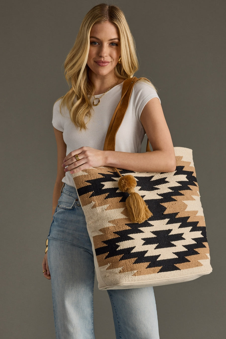 Woman holding a patterned tote bag with a pom-pom detail against a gray background