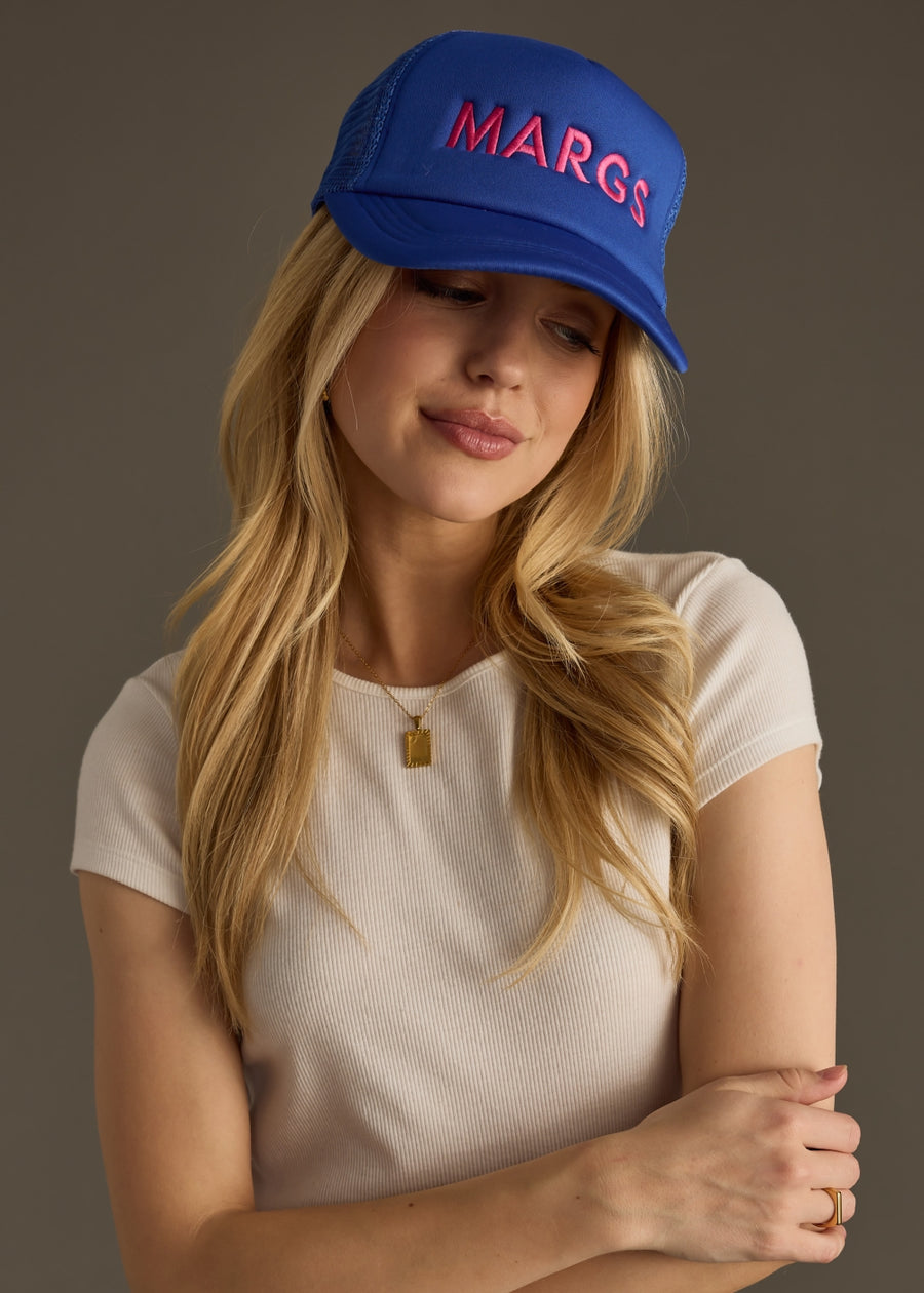 A woman in a fitted white top wears a bright blue baseball cap with bold pink text that reads “MARG.” She’s looking down with a soft smile, arms loosely crossed.