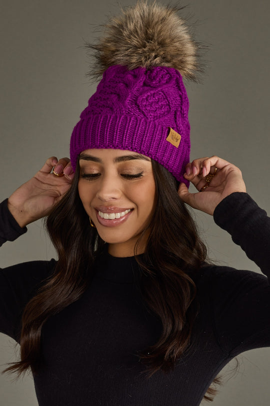Woman wearing a purple knit beanie with a fur pom-pom against a gray background