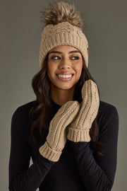Woman wearing a beige knit beanie with pom-pom and matching mittens against a gray background
