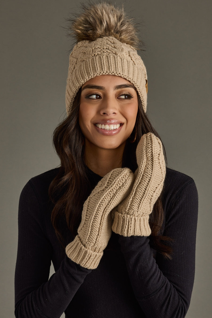 Woman wearing a beige knit beanie with pom-pom and matching mittens against a gray background