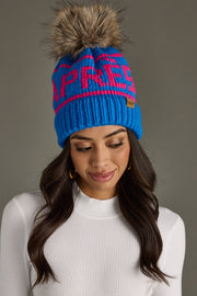 A woman with long brown hair wears Panache Apparel Collection's Hit The Slopes Pom Hat in blue and pink, featuring a faux fur pom and "APRES" lettering, paired with a white ribbed top against a plain gray background.