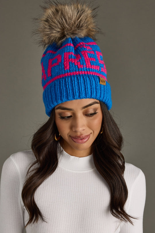 A woman with long brown hair wears Panache Apparel Collection's Hit The Slopes Pom Hat in blue and pink, featuring a faux fur pom and "APRES" lettering, paired with a white ribbed top against a plain gray background.