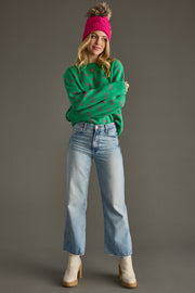 Woman wearing a green sweater with pink patterns, blue jeans, and a pink beanie against a gray background