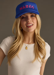 A woman in a fitted white top wears a bright blue baseball cap with bold pink text that reads “MARG.” She’s looking down with a soft smile, arms loosely crossed.