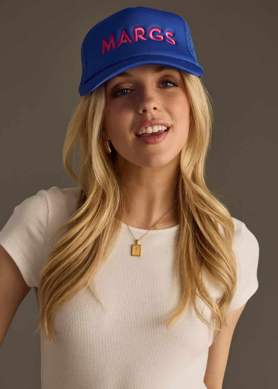 A woman in a fitted white top wears a bright blue baseball cap with bold pink text that reads “MARG.” She’s looking down with a soft smile, arms loosely crossed.