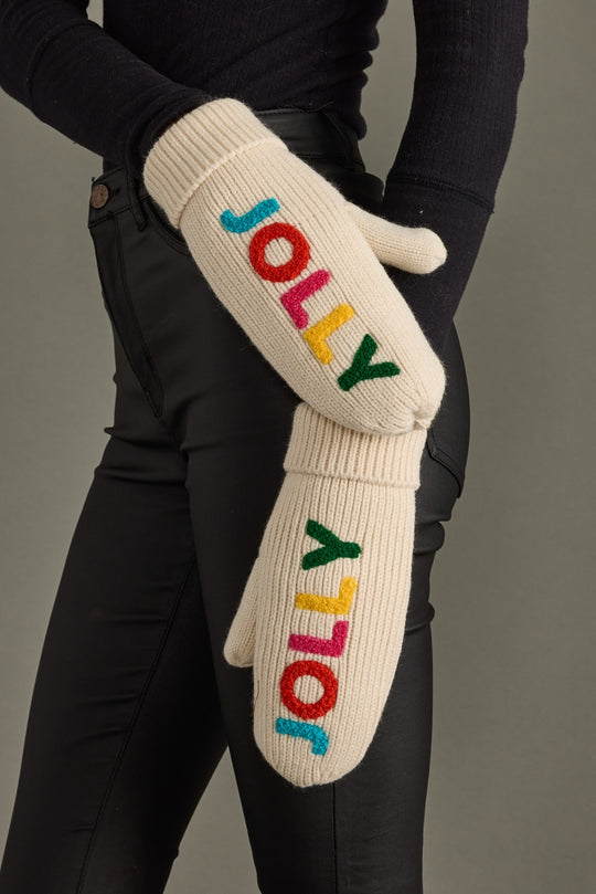 Pair of cream mittens with colorful 'JOLLY' text worn by a person against a gray background