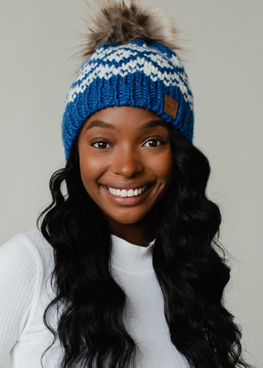 Woman wearing a blue and white patterned knit hat with a fur pom-pom on a plain background