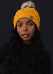 Woman wearing a yellow knit beanie with a fur pom-pom against a black background
