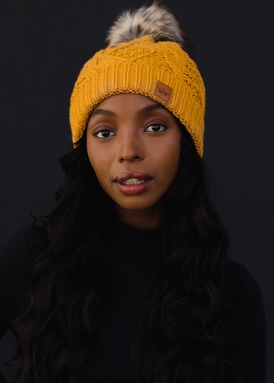 Woman wearing a yellow knit beanie with a fur pom-pom against a black background