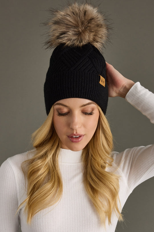 Woman wearing a black knit beanie with a fur pom-pom against a gray background