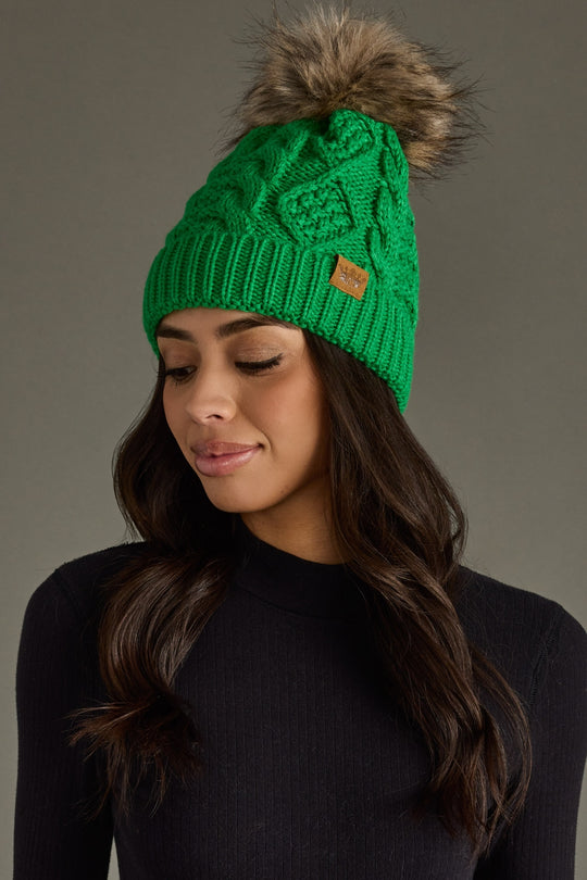 Green knitted beanie with fur pom-pom worn by a model against a gray background