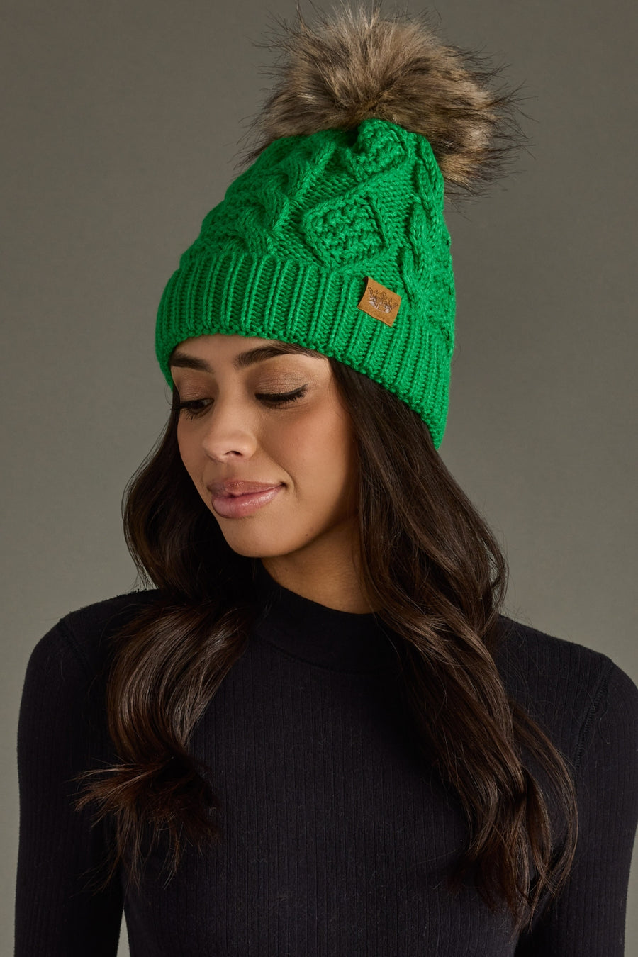 Green knitted beanie with fur pom-pom worn by a model against a gray background