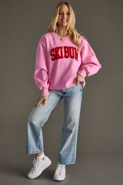 Woman wearing a pink sweatshirt with 'Ski Bum' text, blue jeans, and white sneakers on a gray background
