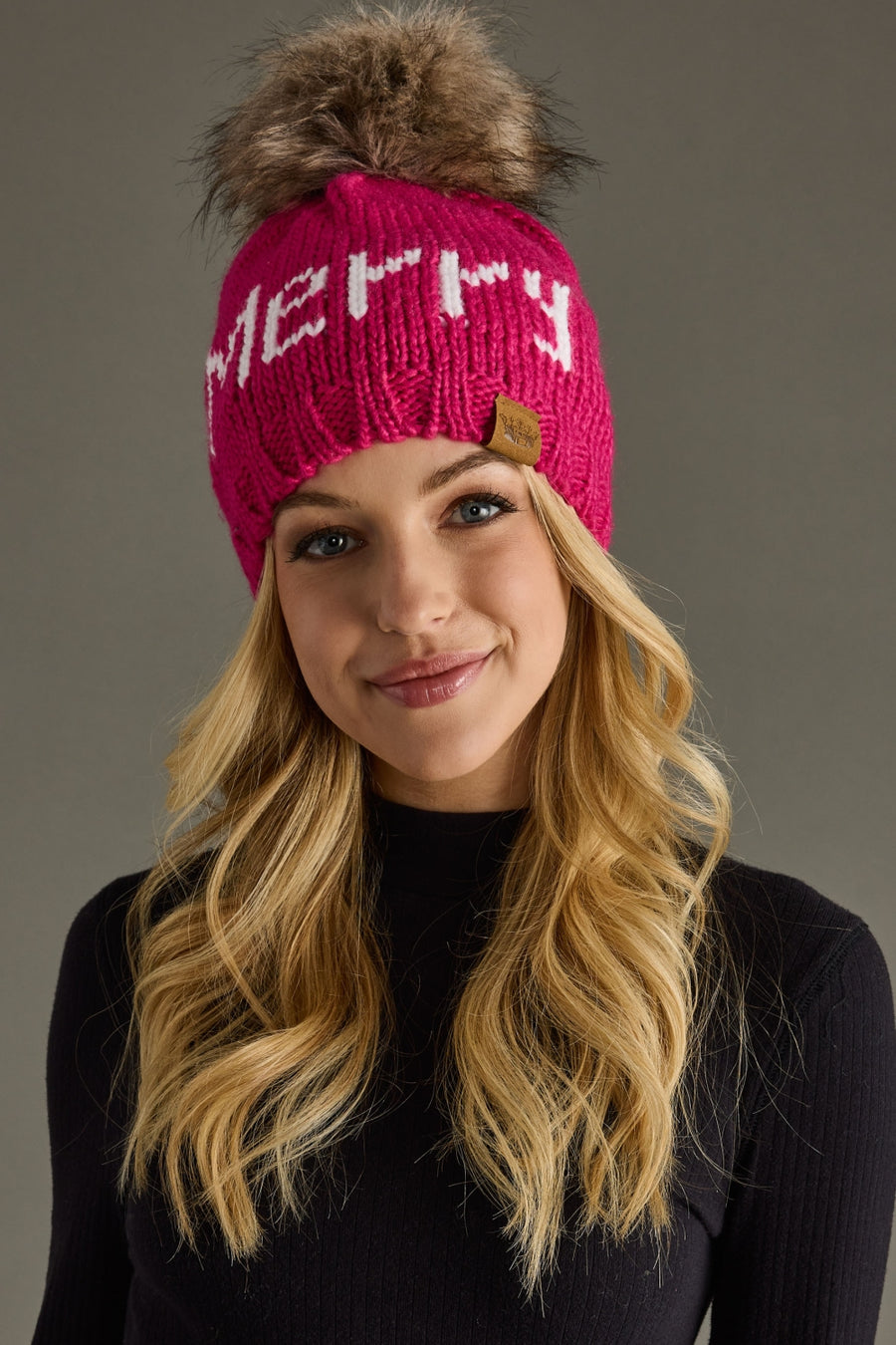 Woman wearing a pink knit hat with 'Merry' text and fur pom-pom against a gray background
