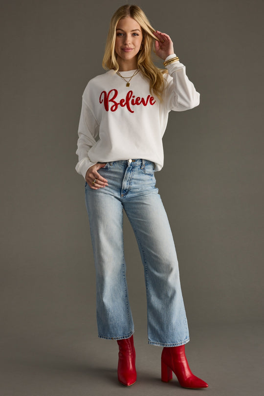 Woman wearing a white sweatshirt with 'Believe' text, blue jeans, and red boots on a gray background