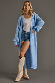 Woman wearing a long light blue cardigan over a white top and denim shorts, standing against a gray background.