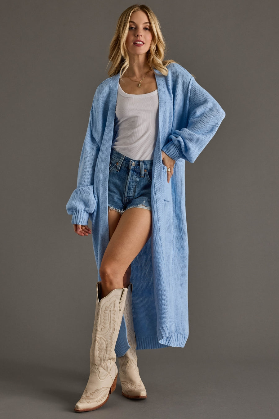 Woman wearing a long light blue cardigan over a white top and denim shorts, standing against a gray background.