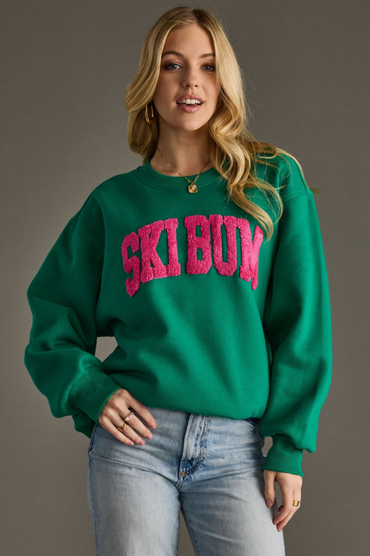 Woman wearing a green sweatshirt with 'Ski Bum' in pink text against a gray background