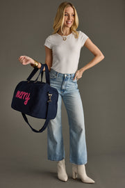 Woman holding a navy duffel bag with 'Merry' embroidered on it, wearing a white t-shirt and blue jeans.