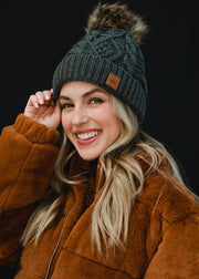A woman with long blonde hair looks directly at the camera with a soft, relaxed expression, wearing a dark gray cable-knit beanie with a faux-fur pom-pom and a small leather logo patch on the front. She’s styled in a black top layered under a warm brown jacket, set against a clean, dark background that emphasizes her natural makeup and cozy fall–winter aesthetic.