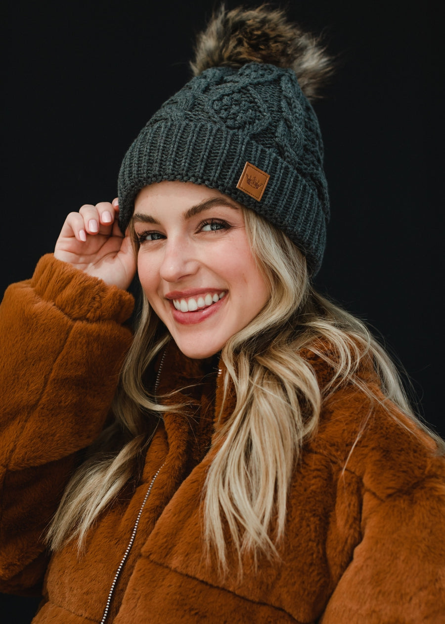 A woman with long blonde hair looks directly at the camera with a soft, relaxed expression, wearing a dark gray cable-knit beanie with a faux-fur pom-pom and a small leather logo patch on the front. She’s styled in a black top layered under a warm brown jacket, set against a clean, dark background that emphasizes her natural makeup and cozy fall–winter aesthetic.