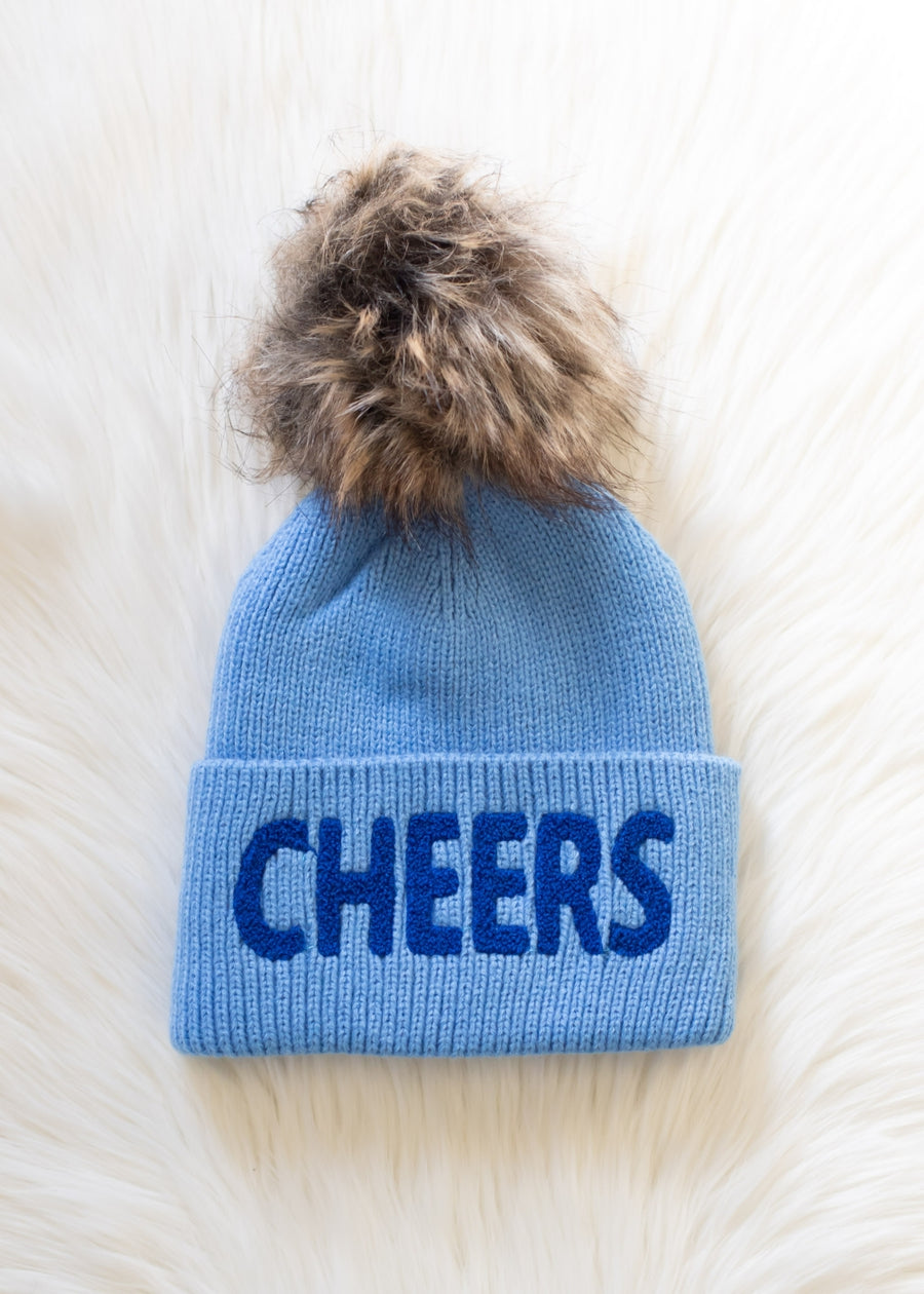 Blue beanie with 'CHEERS' text and fur pom-pom on a light wooden surface
