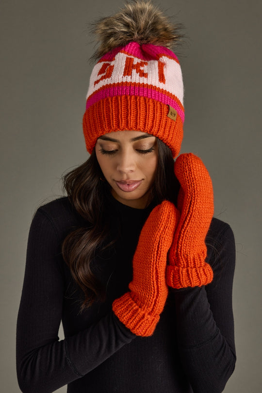 Woman wearing an orange knit hat and mittens with SKI on a gray background