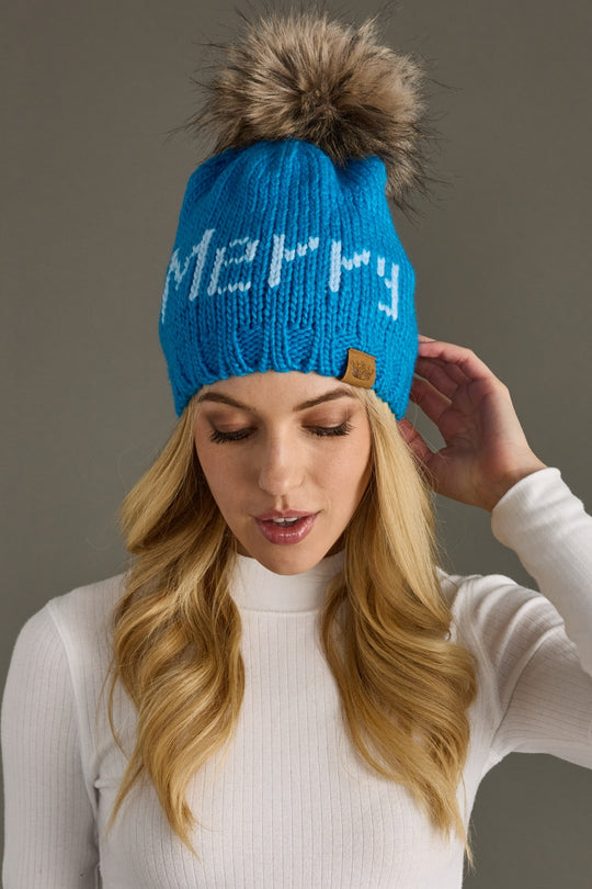 Woman wearing a blue knit beanie with 'Merry' text and fur pom-pom against a gray background