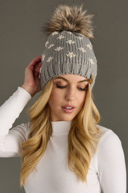 Woman wearing a gray knit beanie with a fur pom-pom against a plain background