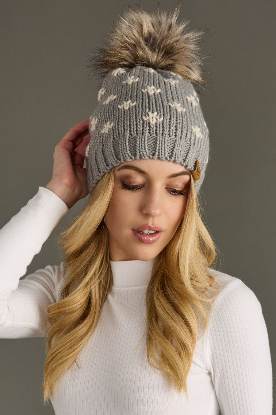 Woman wearing a gray knit beanie with a fur pom-pom against a plain background