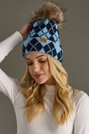 Woman wearing a blue and black checkered beanie with a fur pom-pom against a gray background