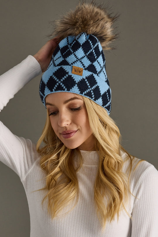 Woman wearing a blue and black checkered beanie with a fur pom-pom against a gray background