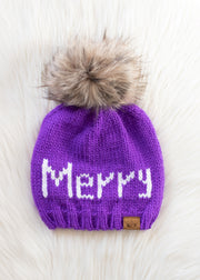 The Merry Pom Hat - Purple by Panache Apparel Collection features a large brown faux fur pom-pom and "Merry" stitched in white yarn, shown on a soft white surface—ideal for pairing with your favorite festive mittens.