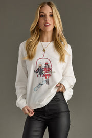 A woman with long blonde hair wears the Alpine Sweatshirt by Panache Apparel Collection, a relaxed fit, lightweight top with an illustration of two people snowboarding. She pairs it with black leather pants and faces the camera against a plain gray background.