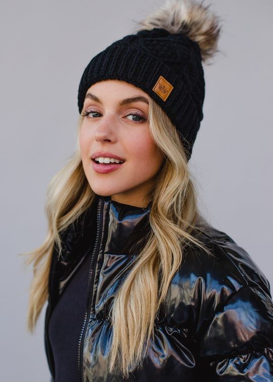 Woman wearing a black knit beanie with a fur pom-pom against a gray background