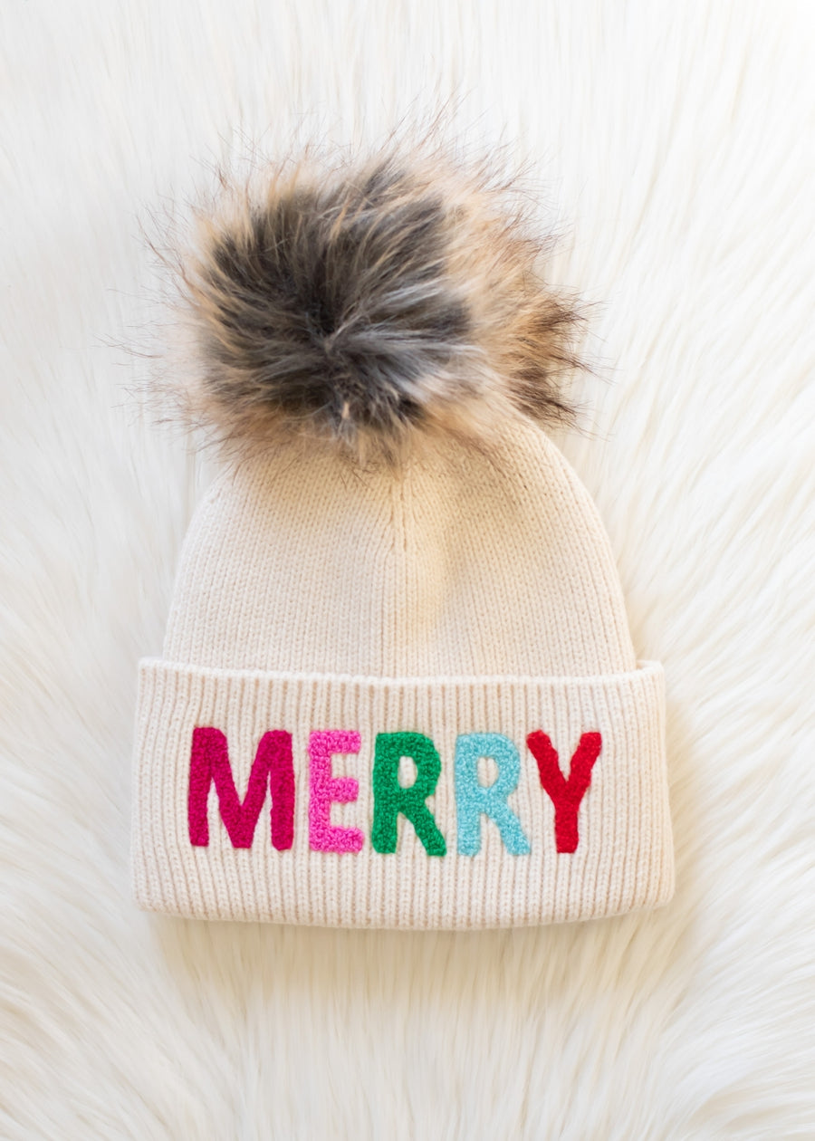 Beige knit beanie with colorful 'MERRY' text and fur pom-pom on a white background