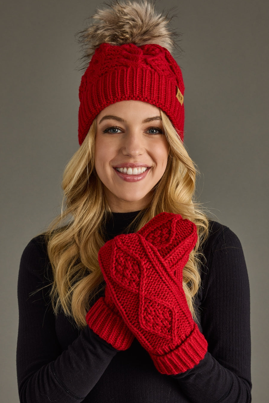 A smiling woman wears the Panache Apparel Collection Red Cable Knit Pom Hat, featuring a faux fur pom, along with matching red knit mittens and a black sweater, standing against a plain grey background.