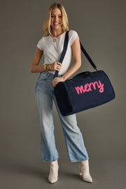 Woman holding a navy duffel bag with 'merry' written on it against a gray background