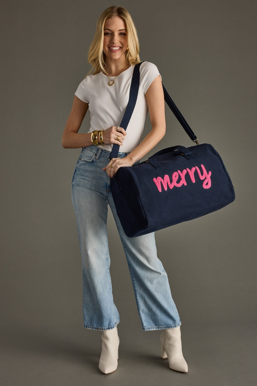 Woman holding a navy duffel bag with 'merry' written on it against a gray background