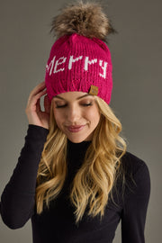 A woman with long blonde hair smiles softly in a black top, wearing the Panache Apparel Collection Merry Pom Hat - Magenta, a fleece-lined knit hat featuring a large faux fur pom-pom and "MERRY" in white text.