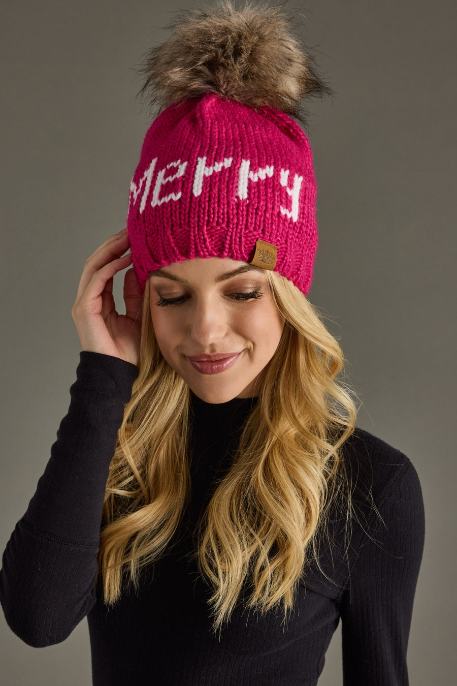 A woman with long blonde hair smiles softly in a black top, wearing the Panache Apparel Collection Merry Pom Hat - Magenta, a fleece-lined knit hat featuring a large faux fur pom-pom and "MERRY" in white text.