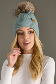 Woman wearing a teal knit beanie with a fur pom-pom against a gray background