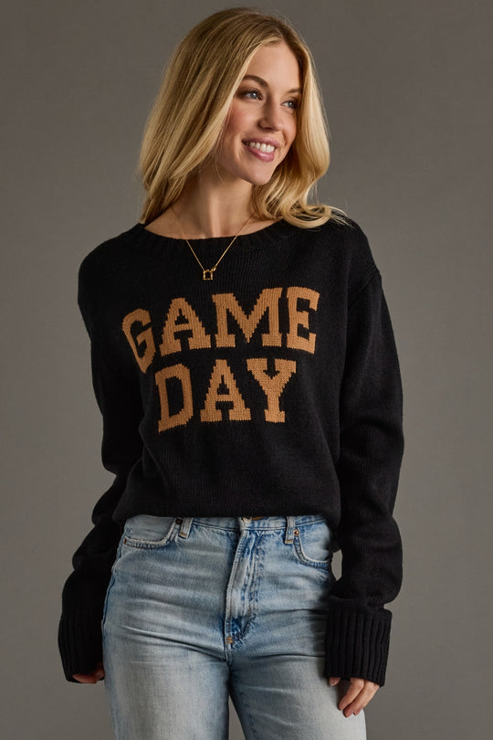 Woman wearing a black sweater with 'GAME DAY' text, standing against a gray background