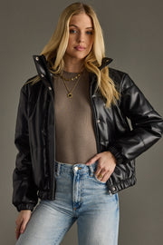 Woman wearing a black leather jacket, brown top, and blue jeans against a gray background