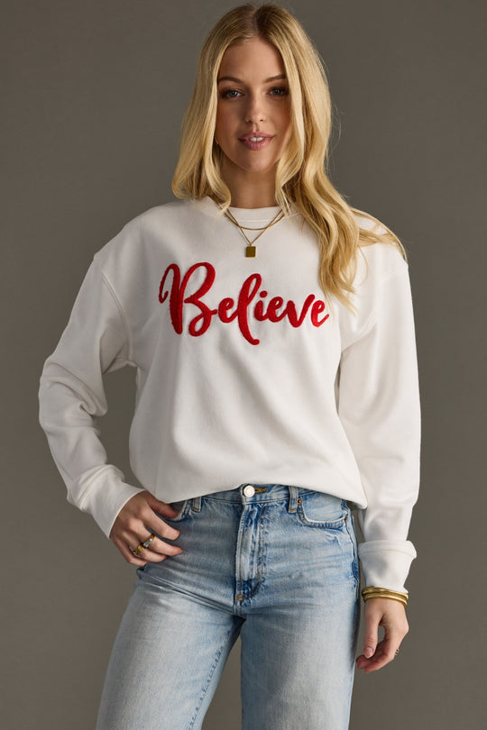 Woman wearing a white sweatshirt with 'Believe' in red text, standing against a plain background.
