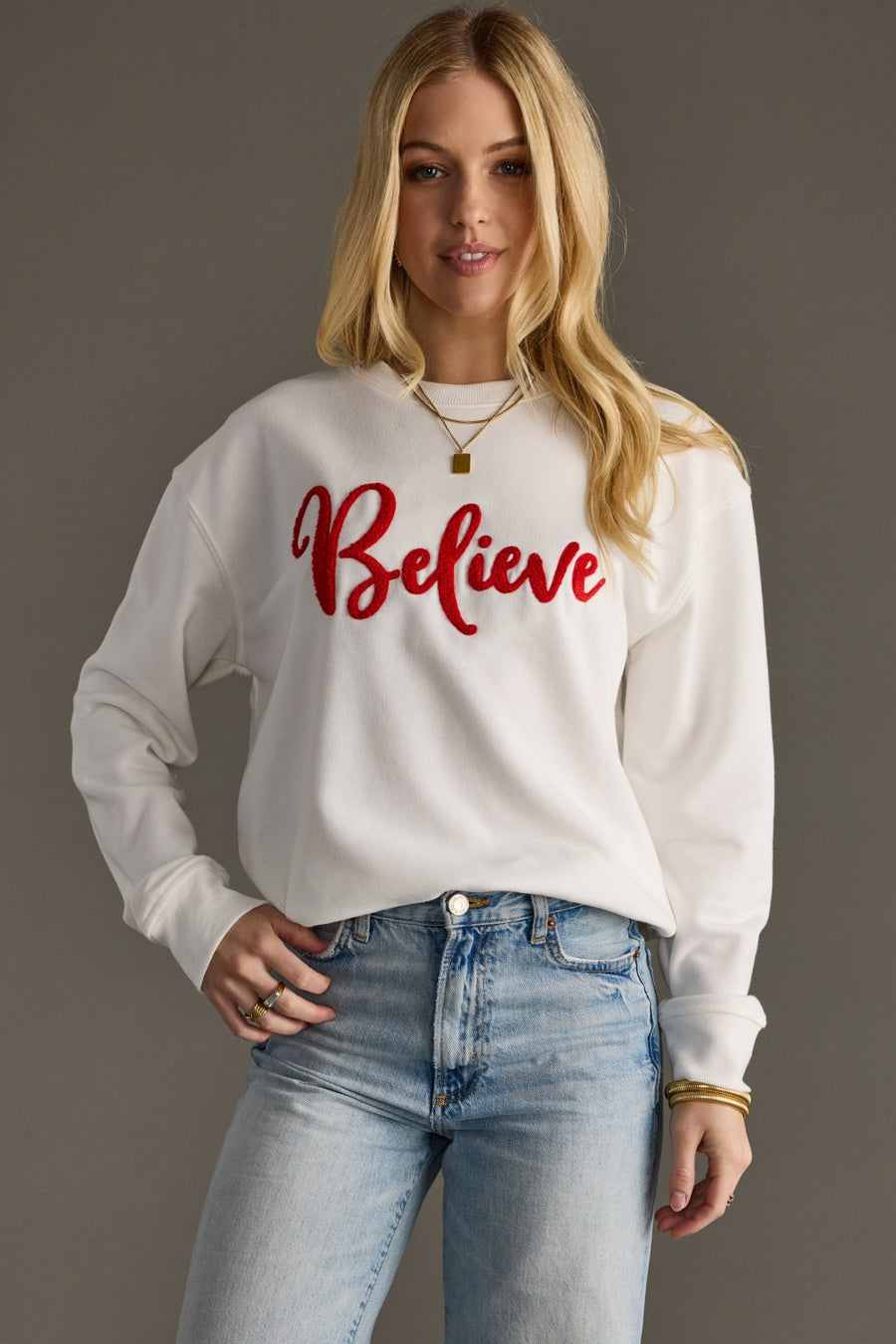 Woman wearing a white sweatshirt with 'Believe' in red text, standing against a plain background.